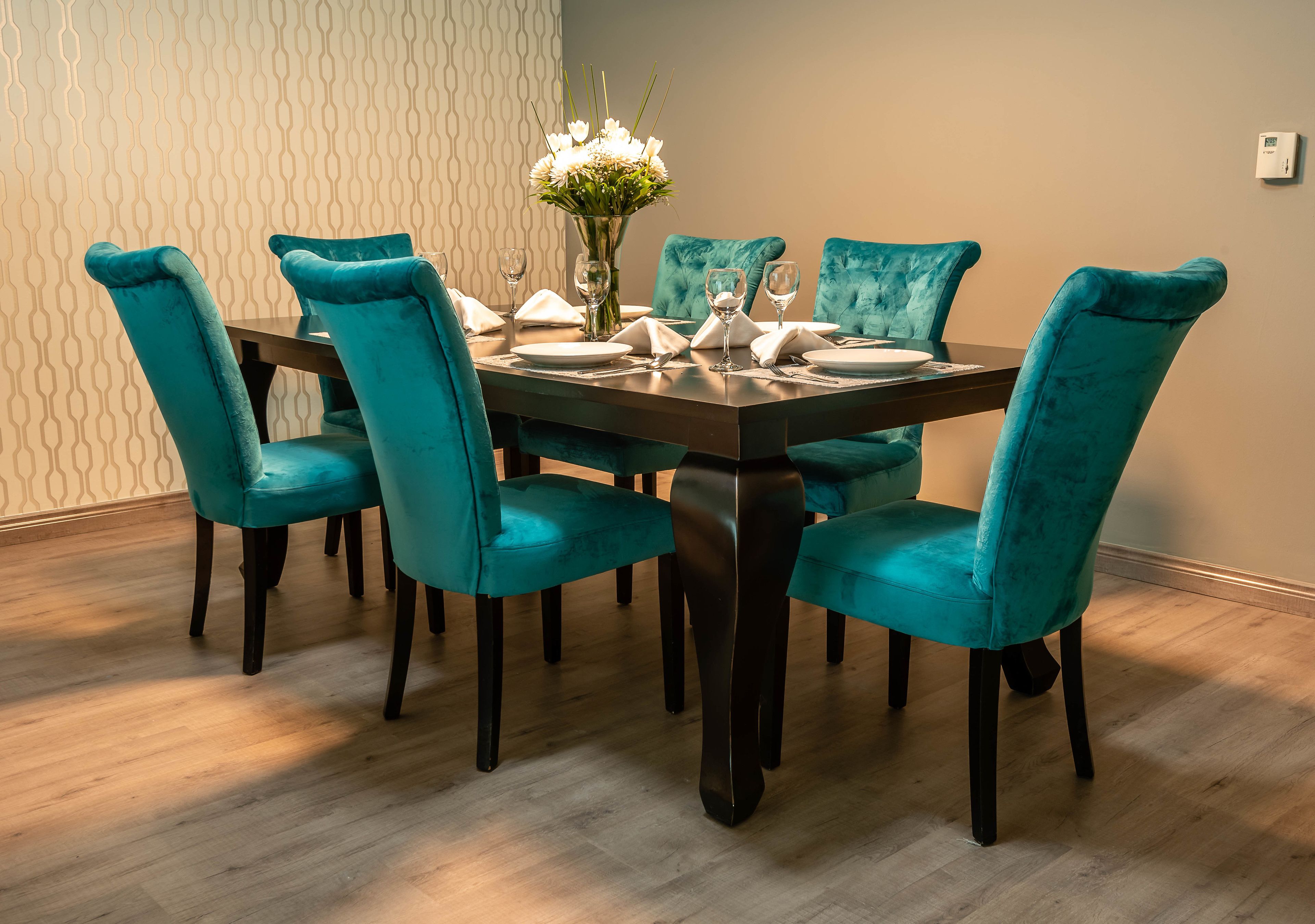 Et spisebord med teal-farvede polstrede stole, en vase med hvide blomster og en trægulv.