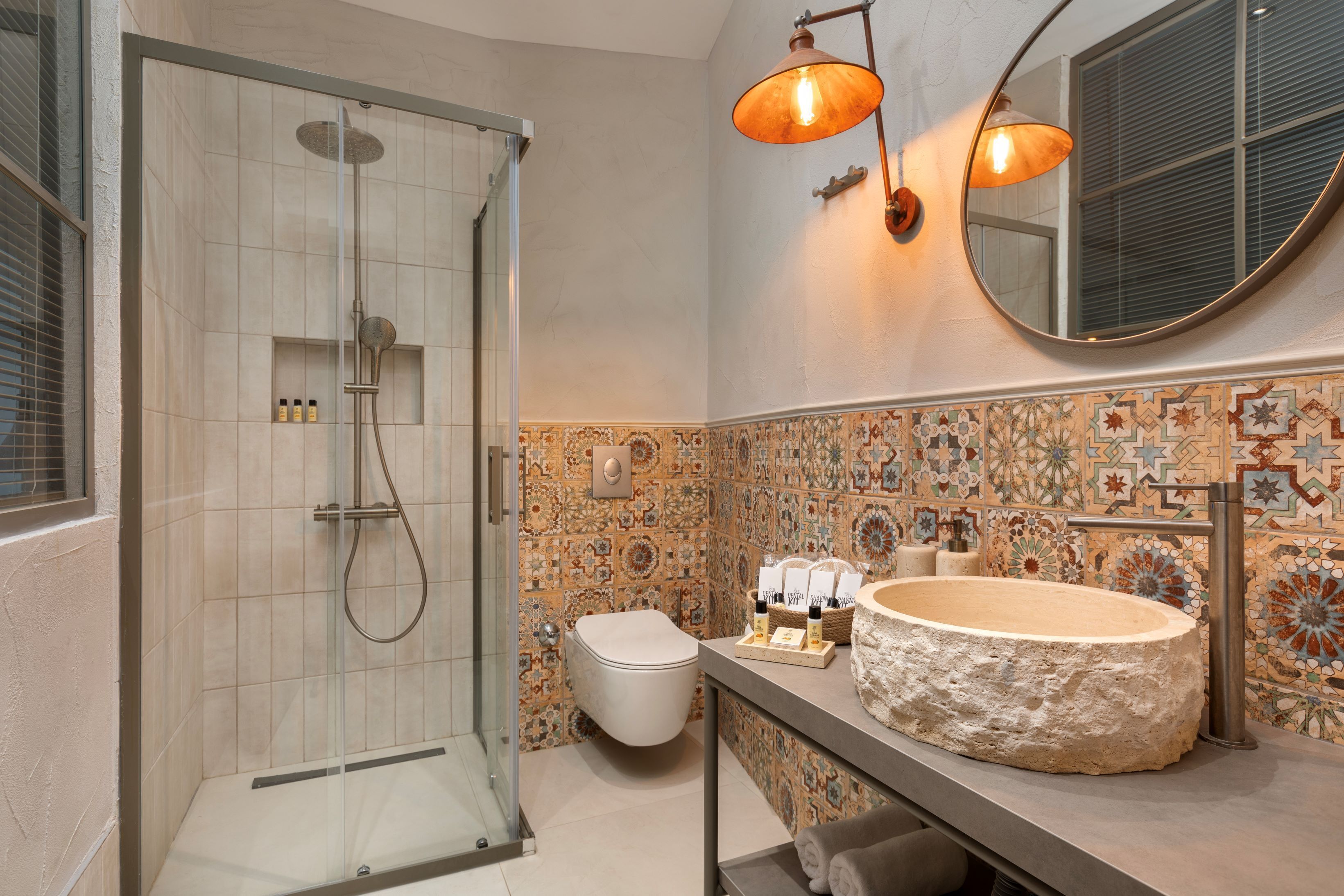 A bathroom with a glass-enclosed shower, a white toilet, a large stone sink, and a tiled backsplash with a floral pattern.