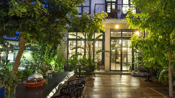 An outdoor patio with wooden flooring, metal chairs, and tables, surrounded by lush greenery and a building with large windows and a balcony. - Casa Sur Antalya (Antalya)