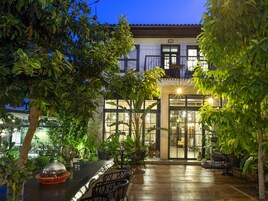 Eine Terrassenanlage im Freien mit Holzboden, Metallstühlen und -tischen, umgeben von üppiger Bepflanzung und einem Gebäude mit großen Fenstern und einem Balkon.