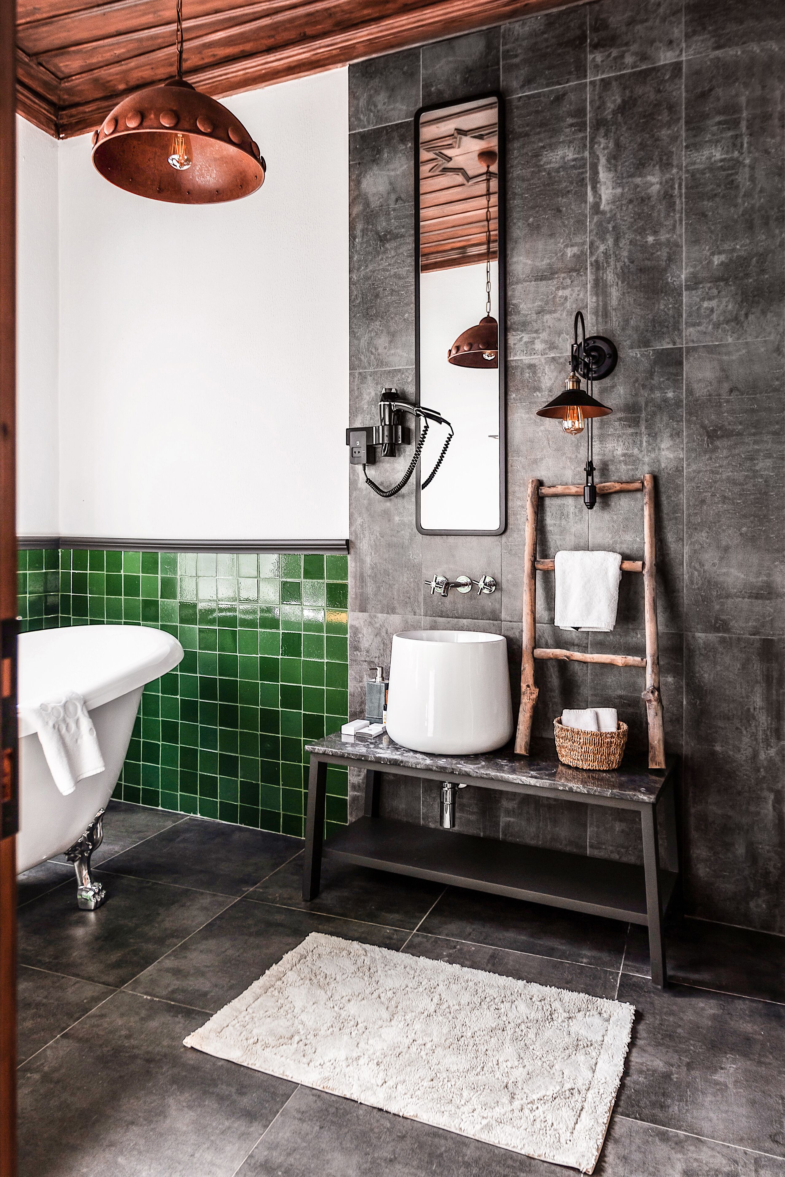 Un baño con una bañera independiente, un espejo grande, un estante escalonado de madera y una lámpara colgante rústica.