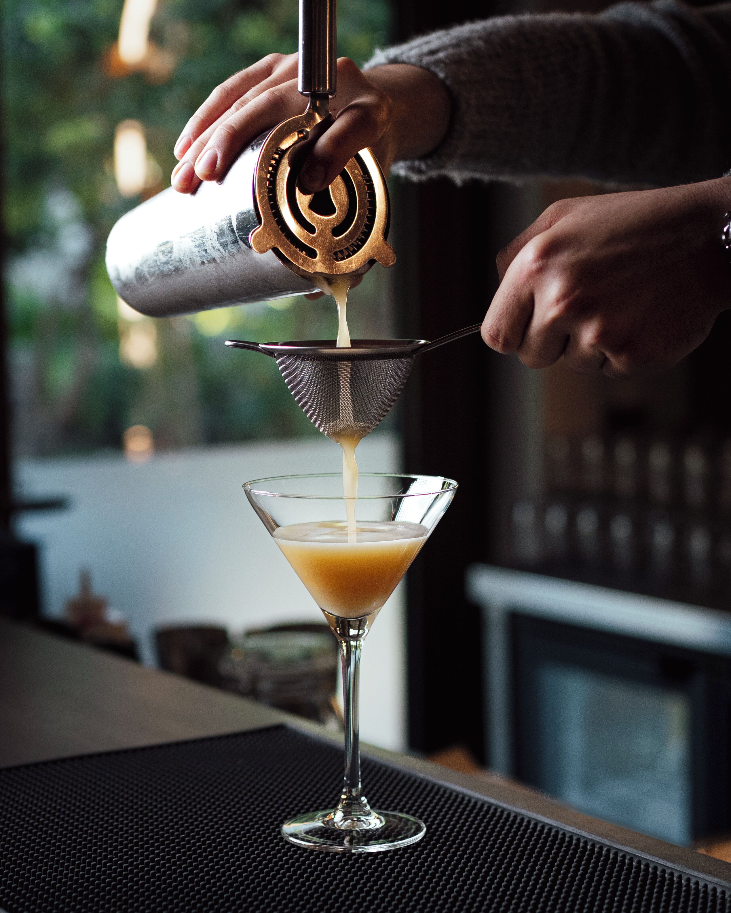 A person pouring a drink into a martini glass using a metal strainer.