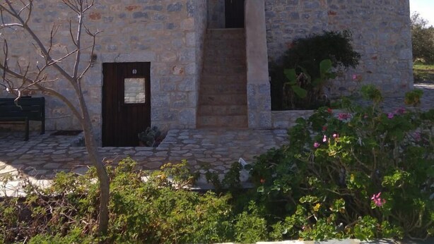 Un bâtiment en pierre avec un escalier menant à une porte, entouré de verdure et d’un mur de pierre.