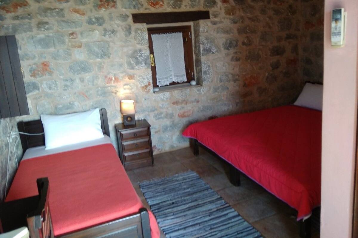 A room with two beds, a stone wall, a small window with a curtain, and a nightstand with a lamp.