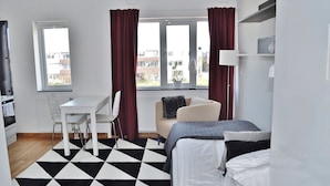 Habitación moderna con cama, escritorio y silla. Cuenta con ventanas con cortinas, un radiador y vistas a edificios.