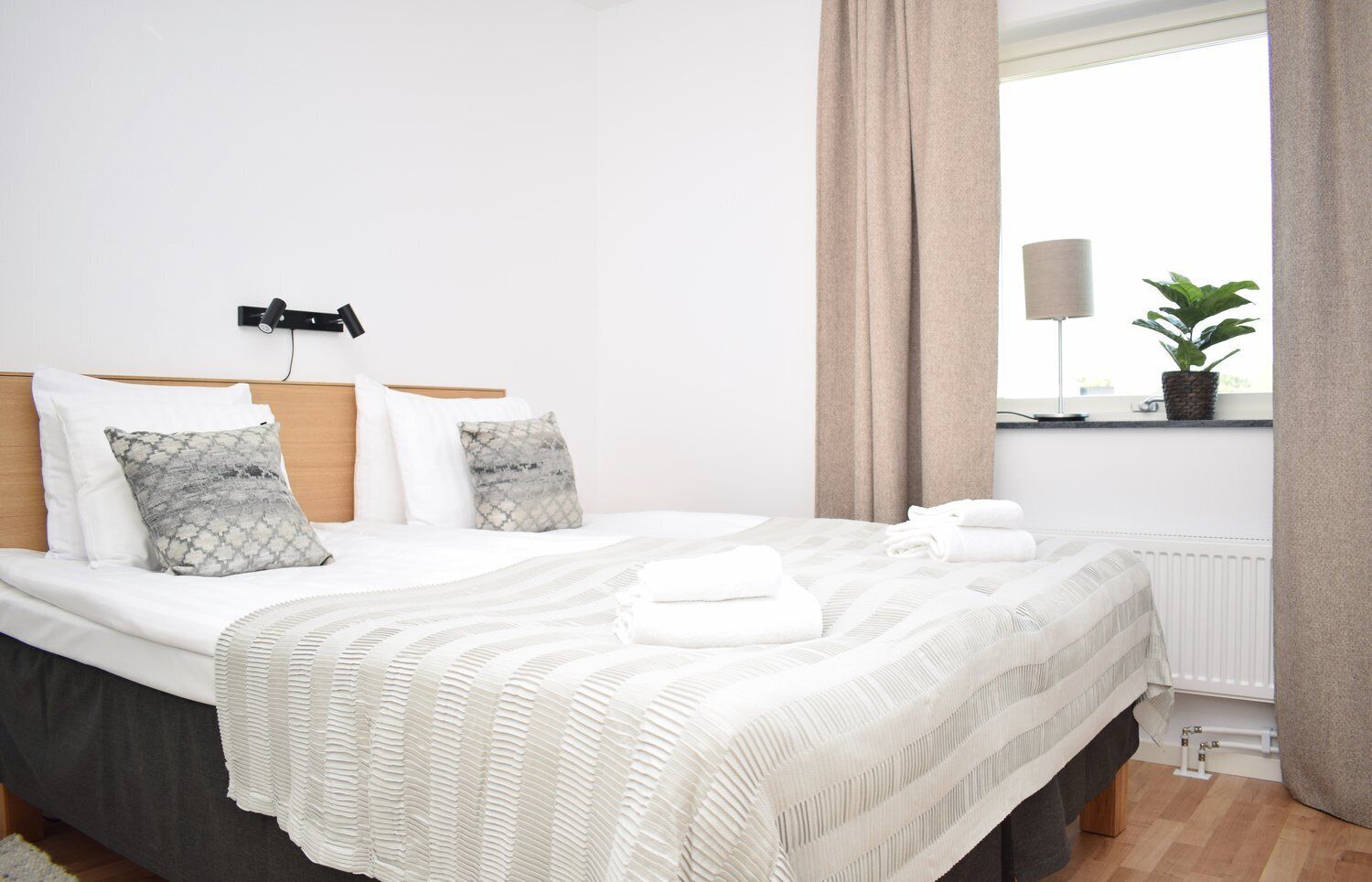 Een net opgemaakt bed met een witte deken en kussens, een bedlampje, en uitzicht op een raam met gordijnen.