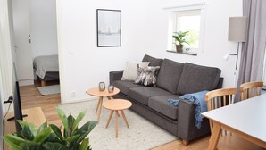 A living room with a grey sofa, two round wooden tables, a TV, a plant, and a window with a plant on the sill. - Företagsbostäder Skarpskyttevägen (Lund)