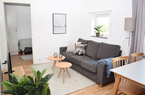 En stue med en grå sofa, to runde træborde, et fjernsyn, en plante og et vindue med en plante i karmen.