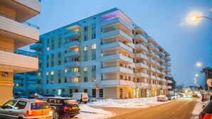 Um edifício de vários andares com varandas, estacionamento e uma rua com veículos.