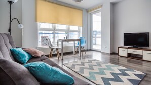 Uma sala de estar moderna com sofá, televisão, mesa de jantar com cadeiras e uma grande janela com persiana amarela.