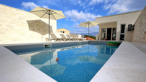 Ein Swimmingpool mit Liegestühlen und Sonnenschirmen, umgeben von einer Steinmauer und einem Gebäude mit großen Fenstern.