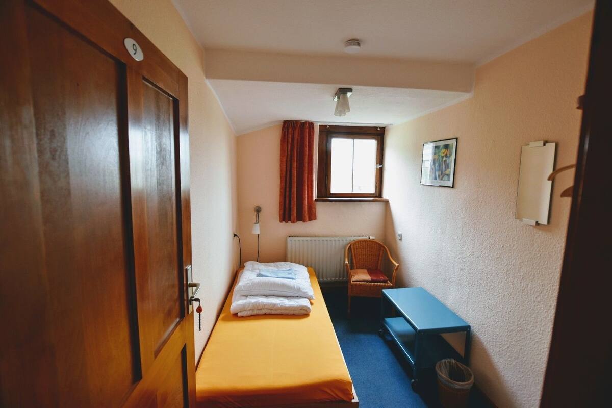 Una habitación pequeña y estrecha con una cama individual, una silla, una mesa y un radiador.