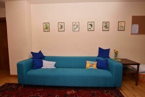 A teal sofa with blue and white cushions, a small table with a vase of flowers, and a bulletin board on the wall. - Herberge Orange - Hostel (Meissen)