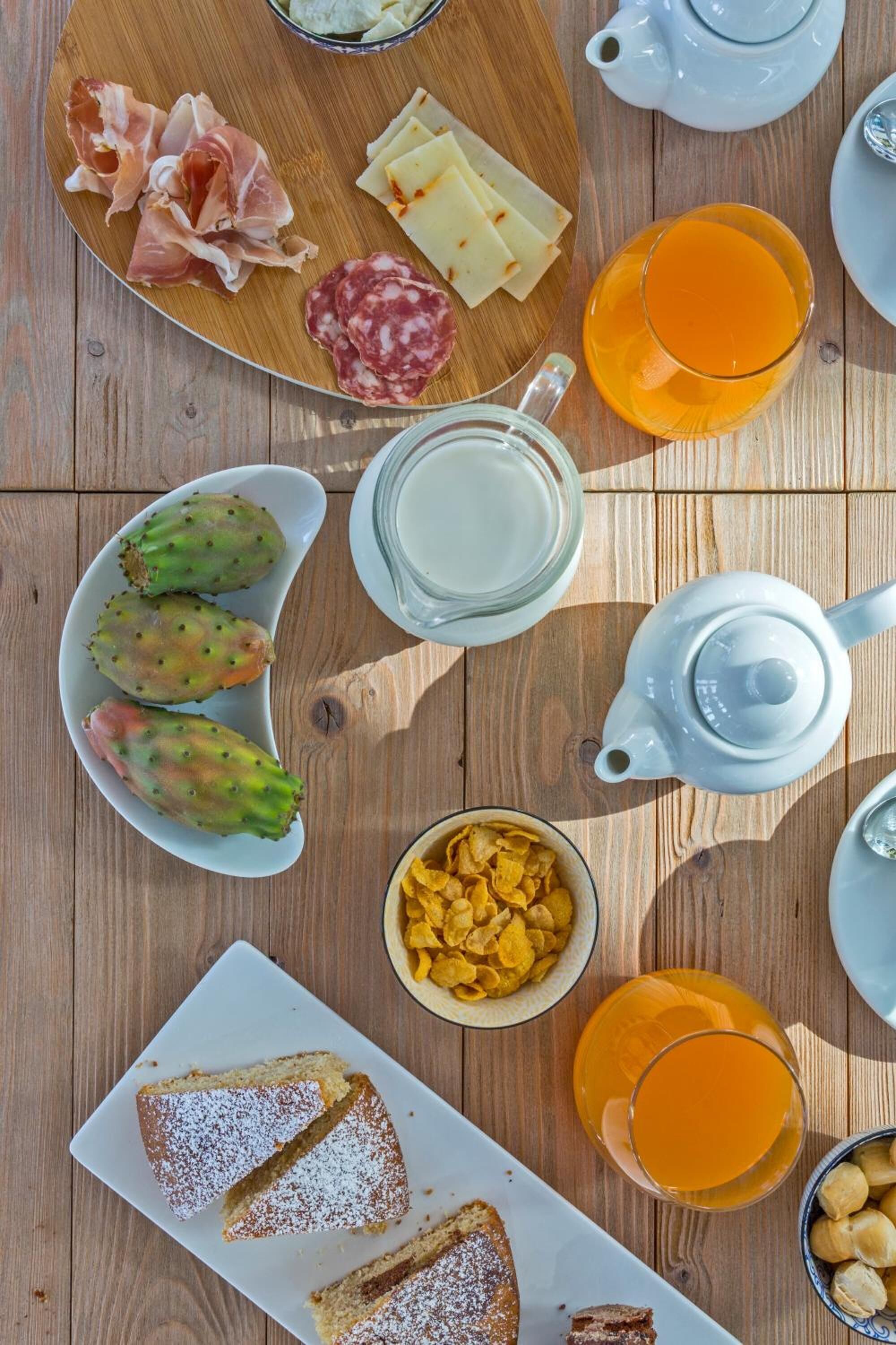 Una mesa de desayuno con diversos alimentos, como queso, embutidos y pan, además de una tetera y tazas de té.