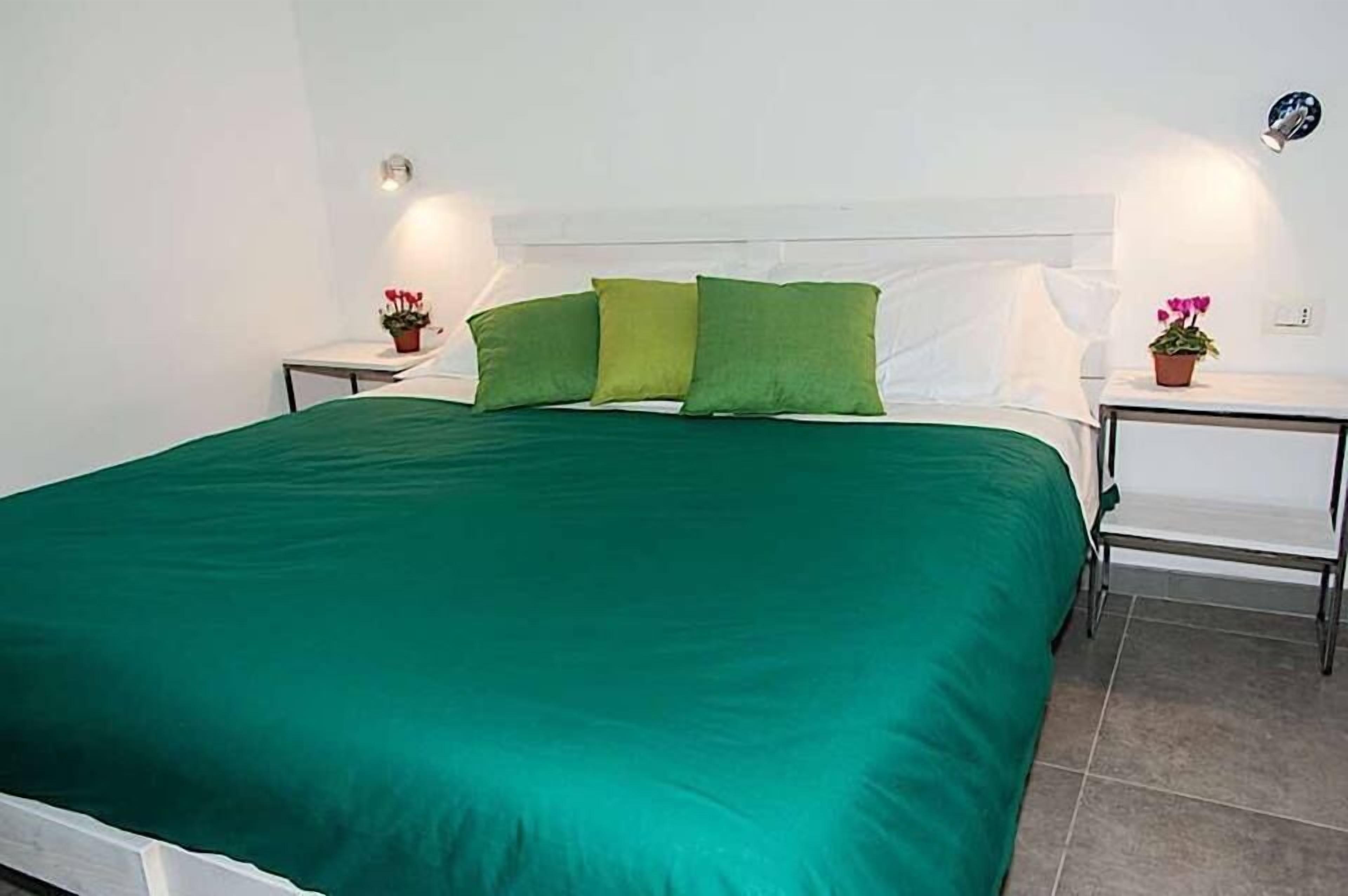 Un lit bien fait, recouvert d’une couvre-lit verte et agrémenté d’oreillers blancs, avec deux tables de chevet de chaque côté, sur lesquelles sont posées de petites plantes en pot.