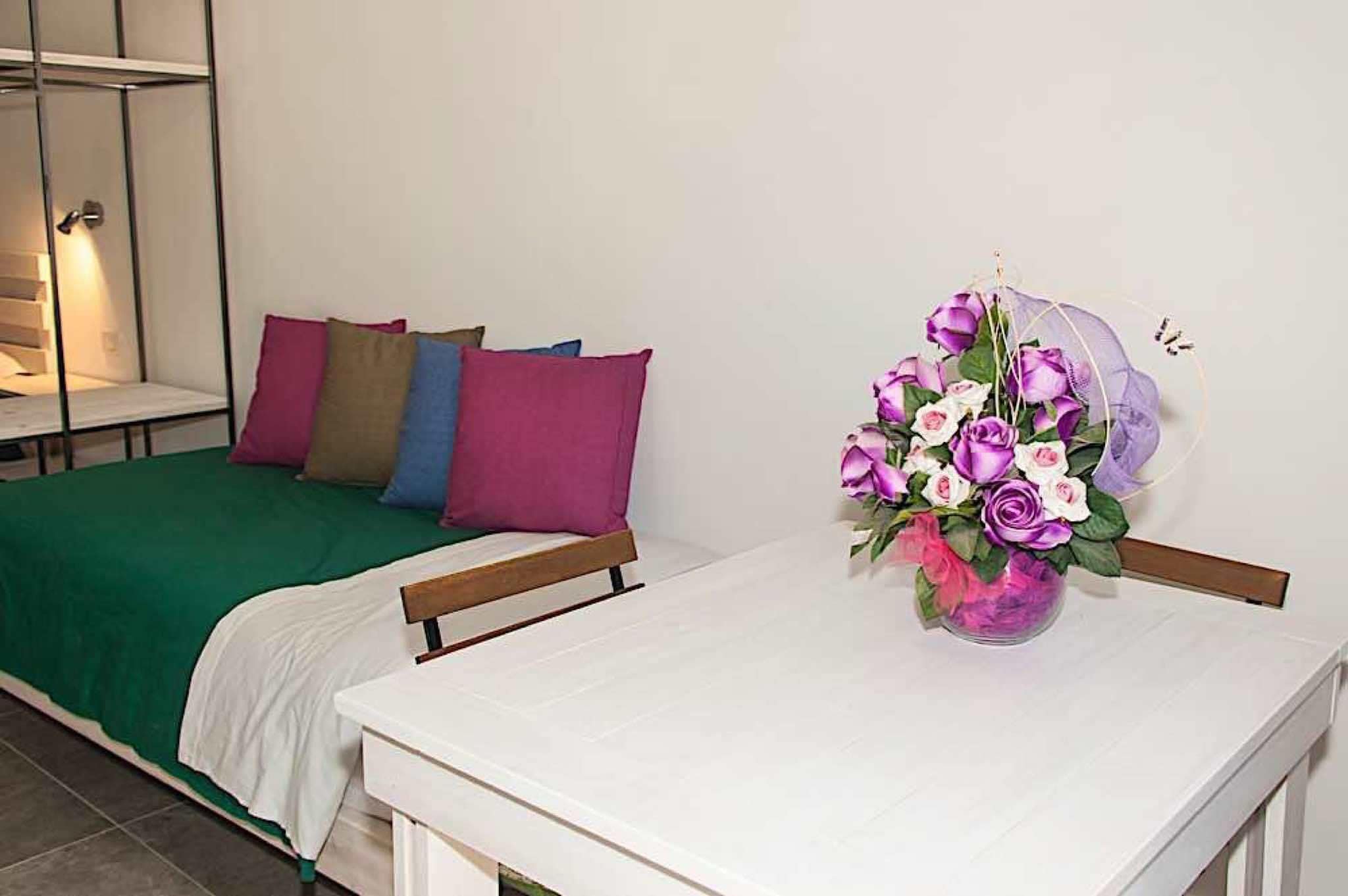 Ein ordentlich bezogenes Bett mit einer grünen Tagesdecke und mehreren Kissen in verschiedenen Farben. Neben dem Bett steht ein weißer Tisch mit einer Vase voller lila und weißer Blumen.