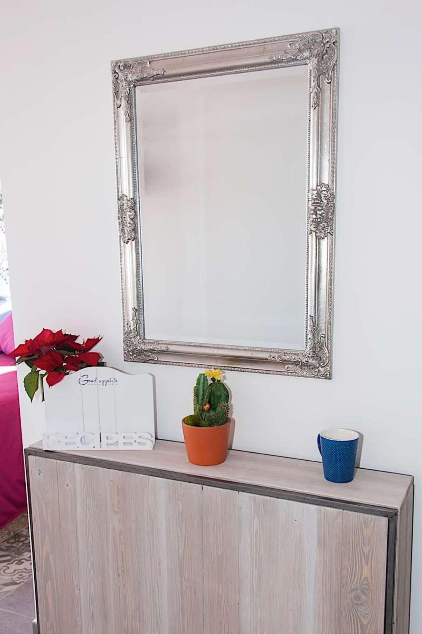 Un espejo montado en la pared con un marco decorativo, una pequeña maceta, un cartel con la palabra "RECIPES" y una taza azul sobre un mueble de madera.
