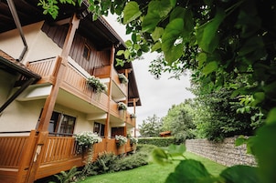Un edificio de madera de dos pisos con balcones, rodeado de vegetación y un muro de piedra.