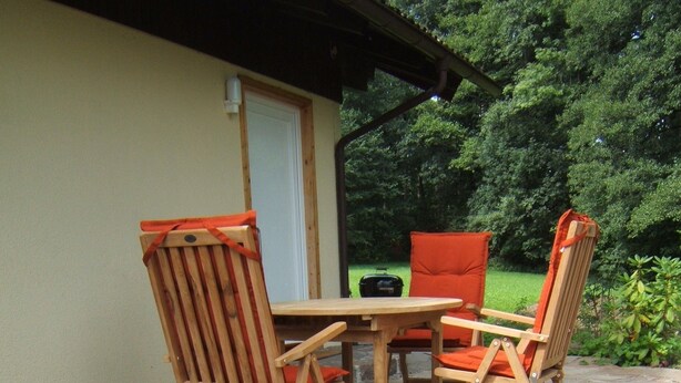 An outdoor patio with a wooden table and chairs, surrounded by greenery and flowers. - Ferienwohnung Schwahn (Herdwangen Schoenach)