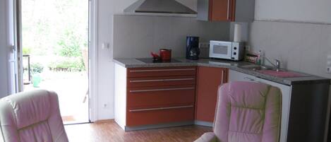 A compact kitchen with a microwave, coffee maker, and cabinets. There are two chairs with cushions, a small table, and a sliding glass door leading to an outdoor area.