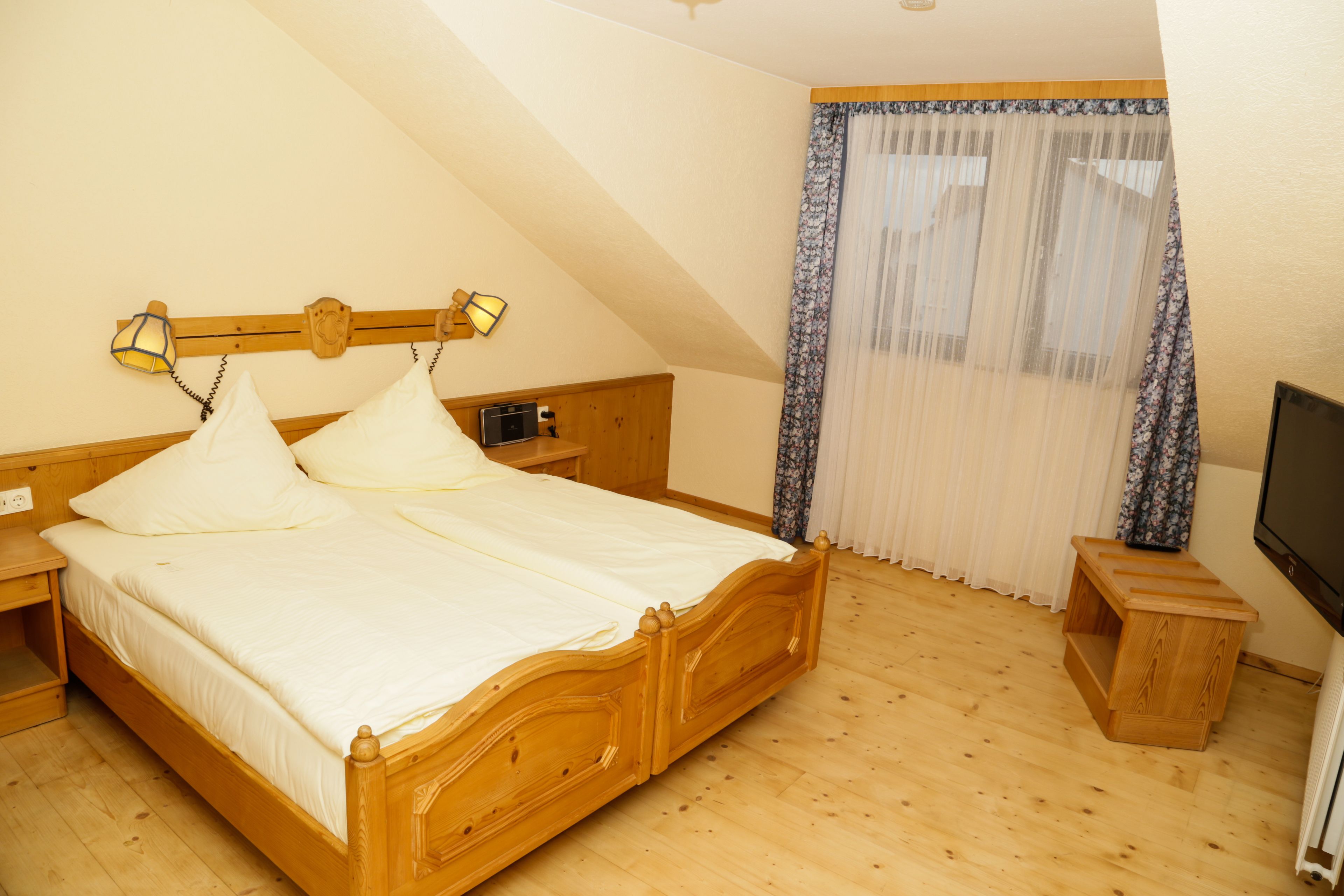 A bedroom with a wooden bed, a nightstand, a bedside table, a TV, and a window with sheer curtains.