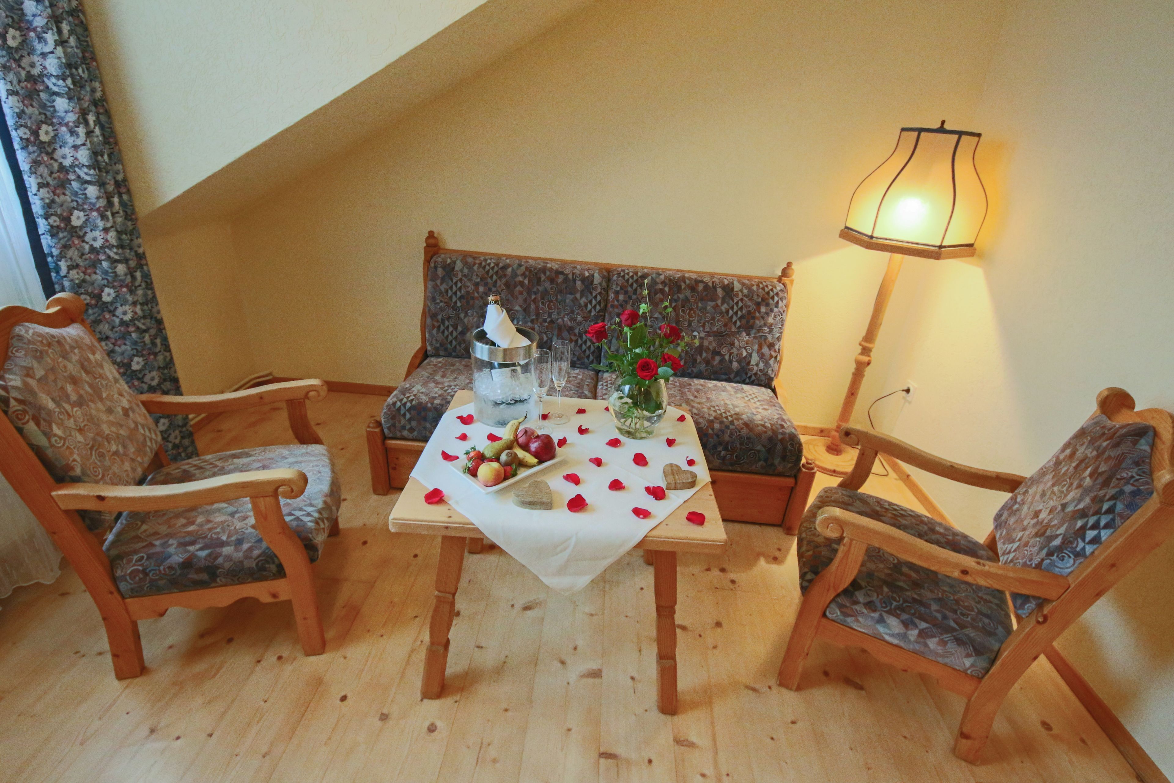 Un rincón acogedor con un sofá y dos sillas de madera, una mesa con mantel blanco, un florero con flores rojas y una lámpara de mesa.