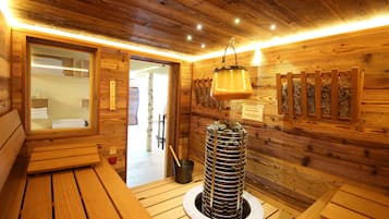 A wooden sauna room with benches, a central stone structure, and a hanging light fixture.