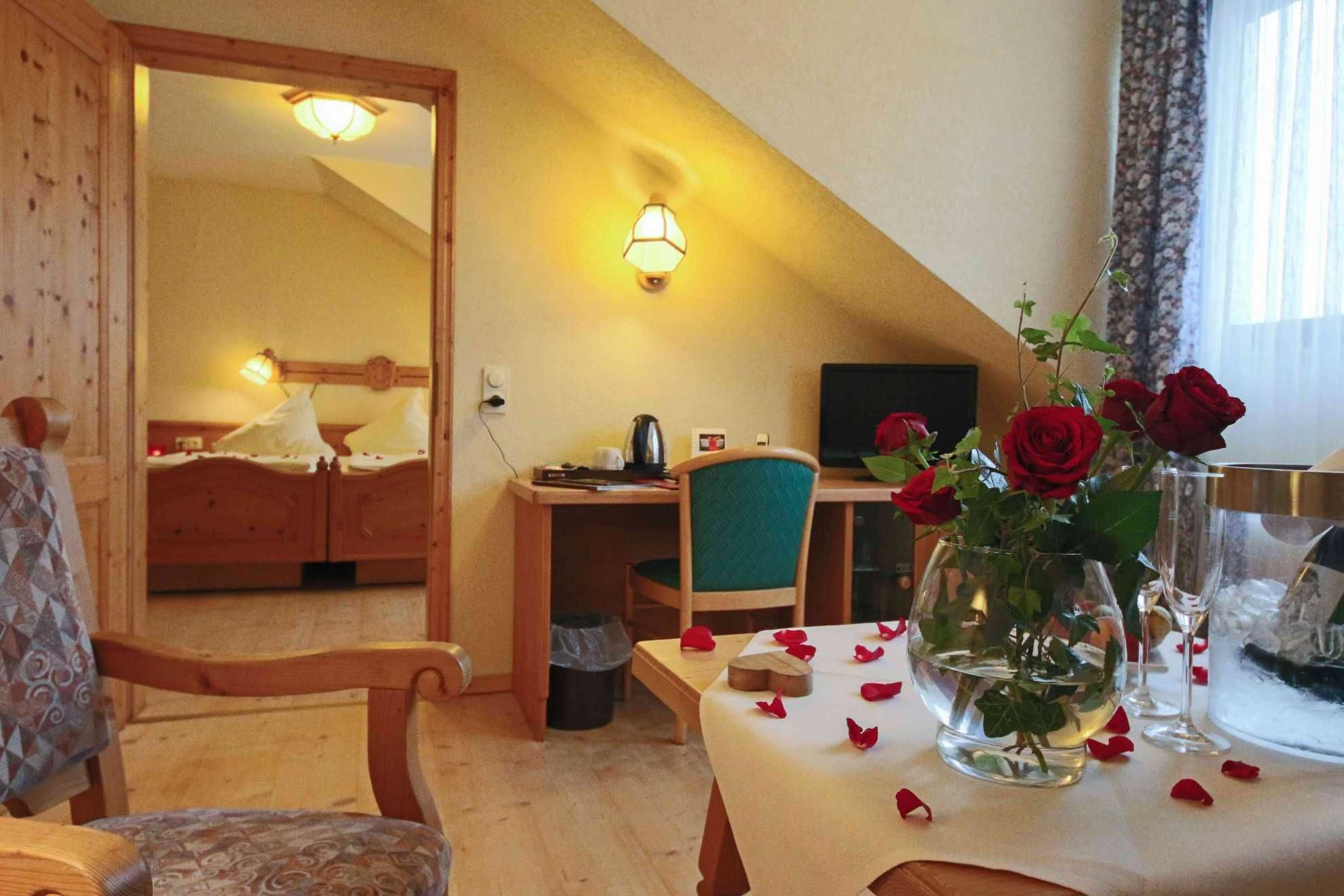 Une chambre d’hôtel avec un lit, un bureau, une chaise et une table décorée de pétales de rose et agrémentée d’un vase rempli de roses.