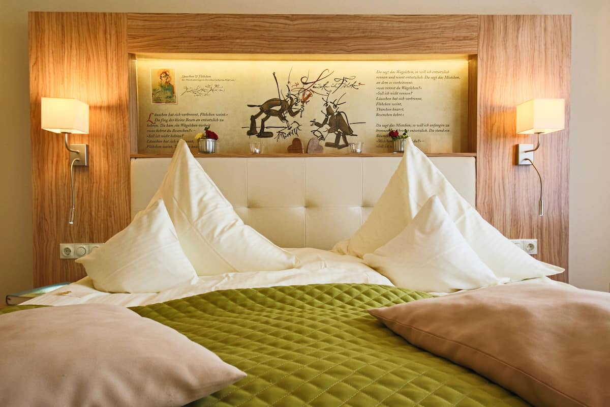 A hotel room with a large bed, a green quilted bedspread, and a headboard with a framed artwork. There are two wall-mounted lamps on either side of the bed.