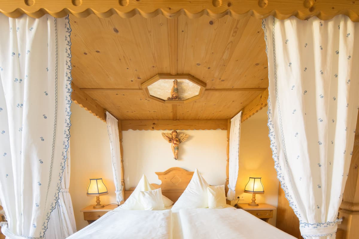 A wooden ceiling with a hexagonal skylight, a bed with white linens and two bedside lamps, and a wooden headboard with a cross.