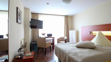 Quarto de hotel com cama, escrivaninha com cadeira, televisão e janela com cortinas.