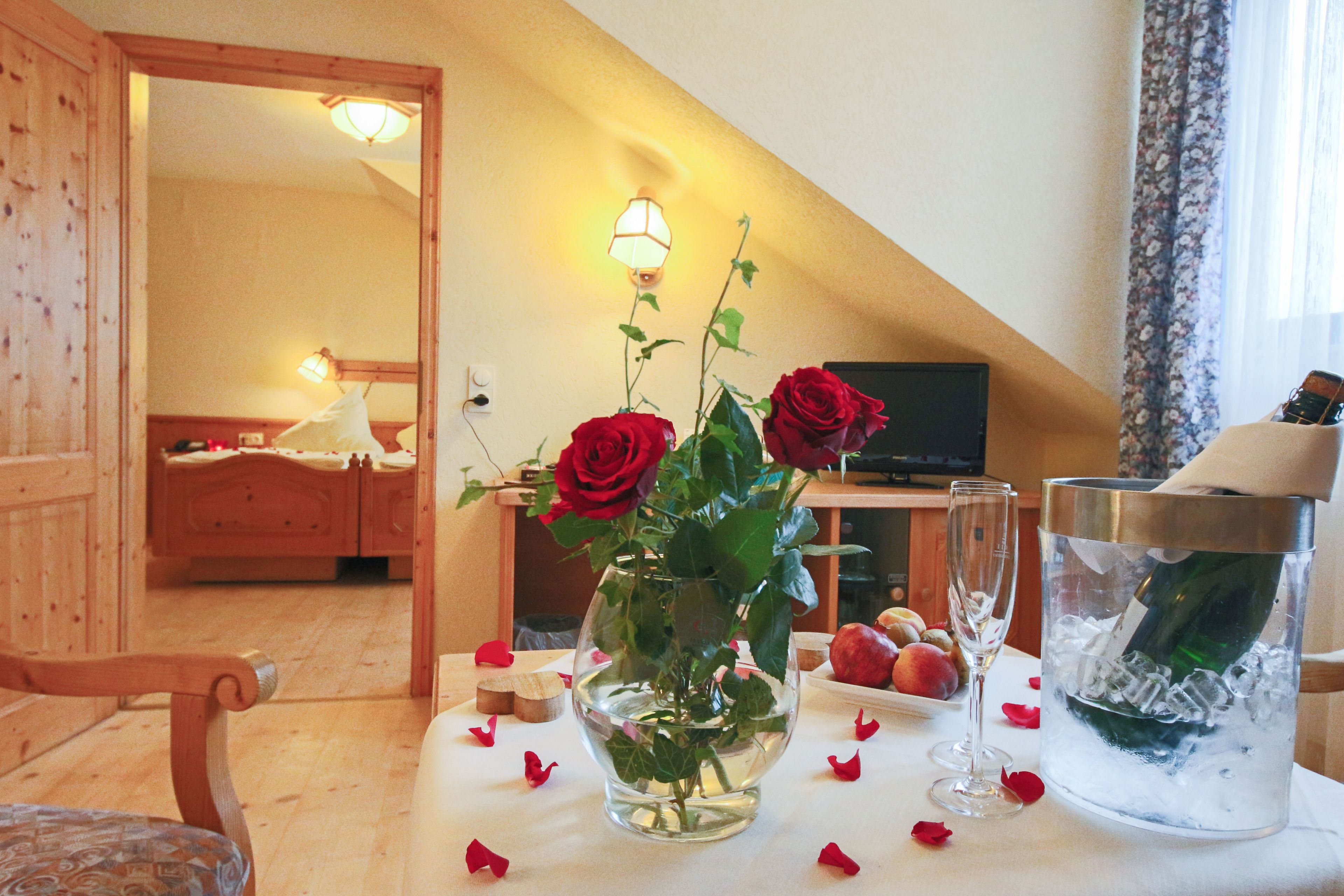 Une chambre avec un lit en bois, une petite table sur laquelle est posé un vase de roses rouges, une bouteille de champagne et un verre.