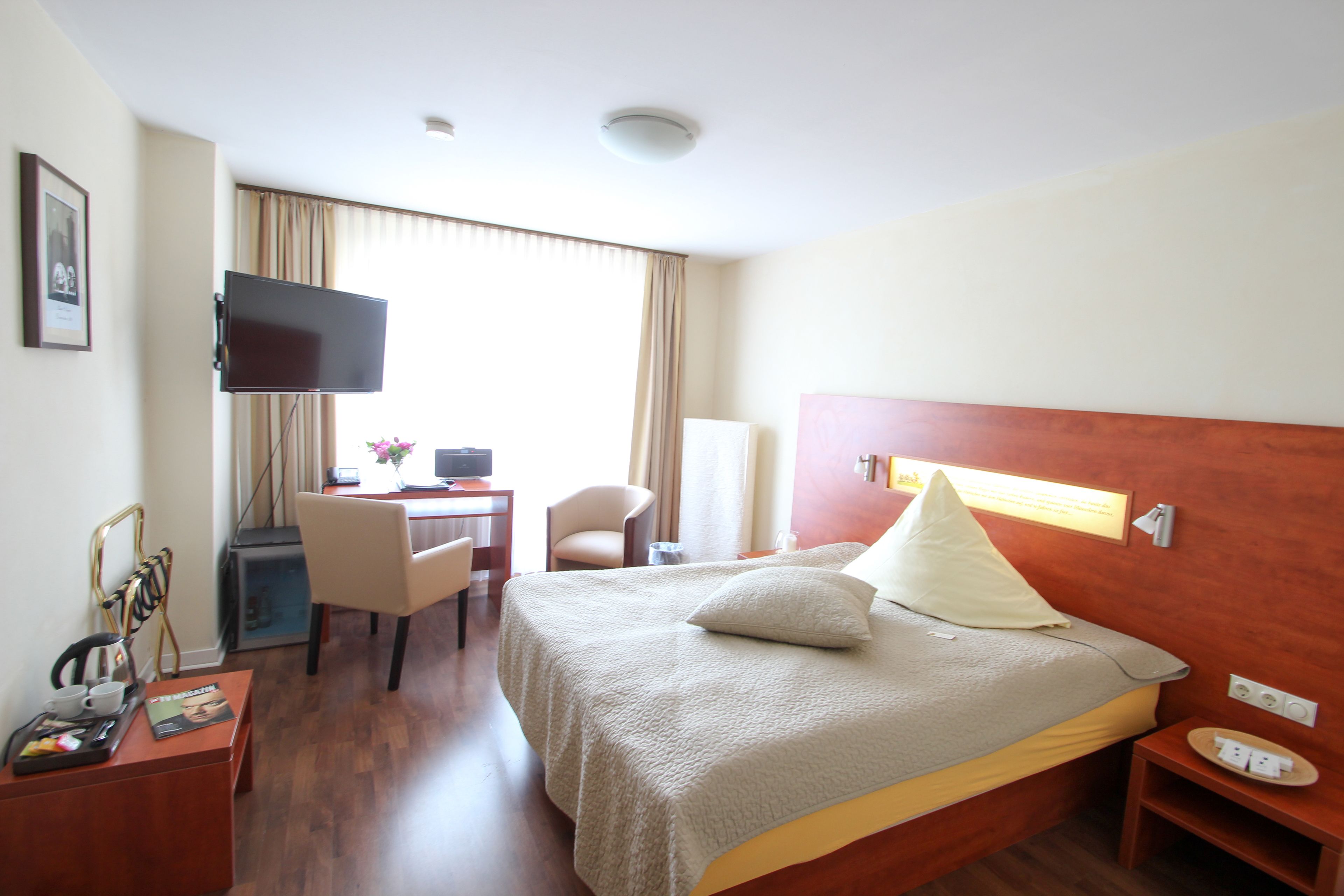 A hotel room with a bed, a desk with a computer, a chair, a TV mounted on the wall, and a small table with a tray.
