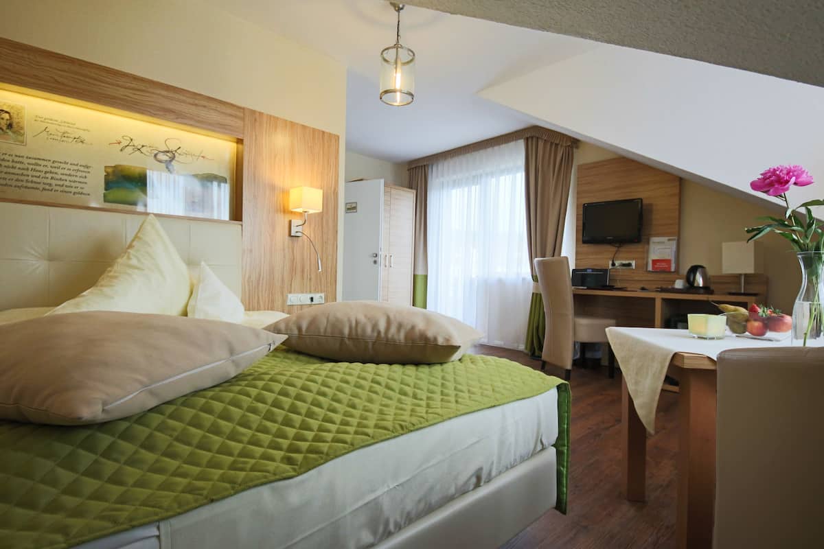 A hotel room with a bed, a desk, a chair, a table, a television, and a vase with flowers.