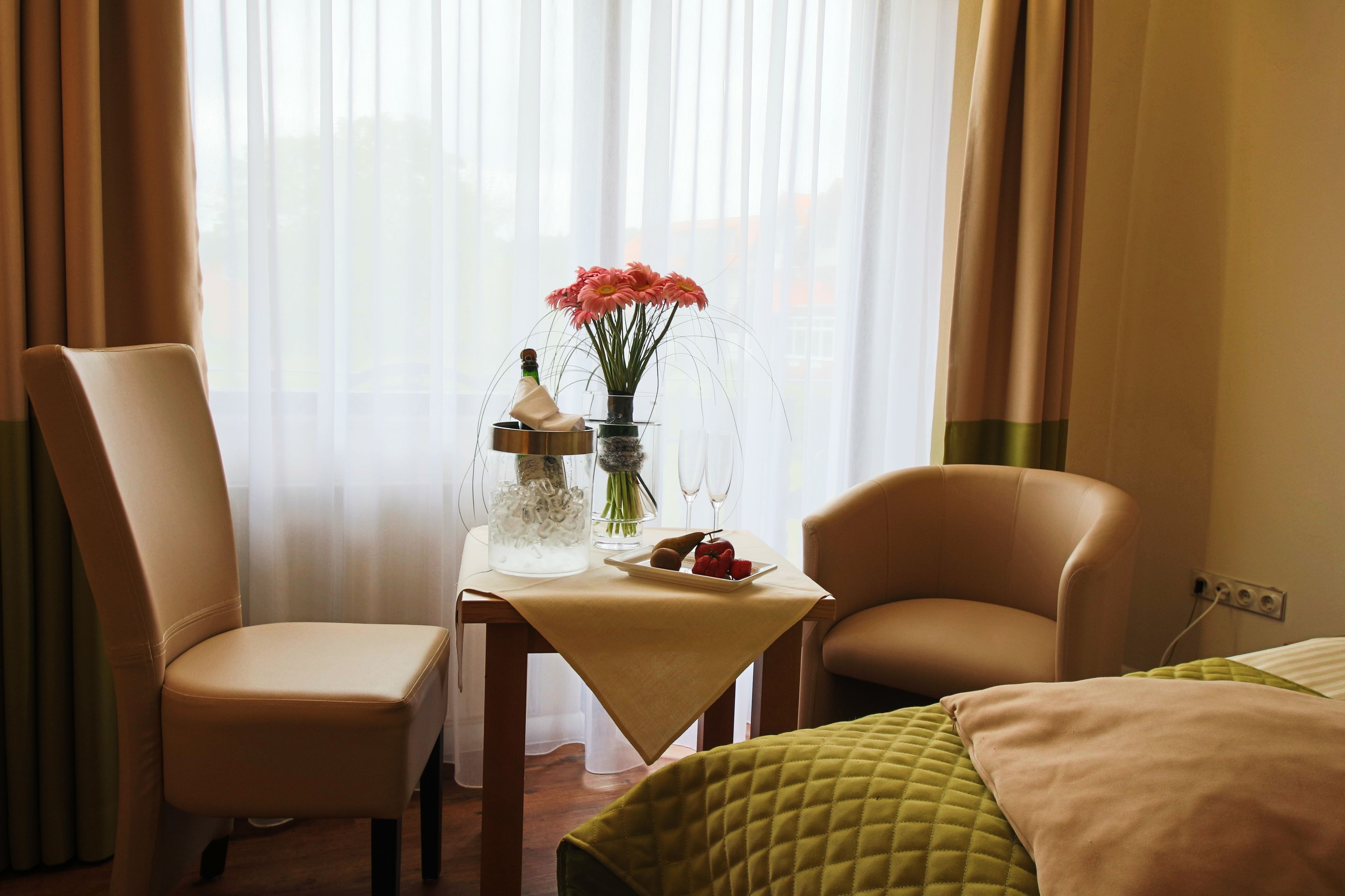 A hotel room with a bed, two chairs, a table with a vase of flowers, and a plate of fruit.
