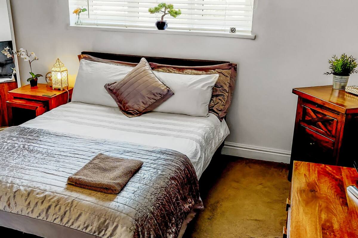 A neatly arranged bedroom with a bed, bedside tables, a TV, and a window with plants.