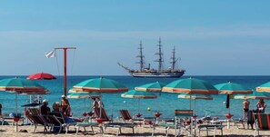 Una playa con hileras de sombrillas y sillas de descanso, un barco grande al fondo y cielos azules despejados.