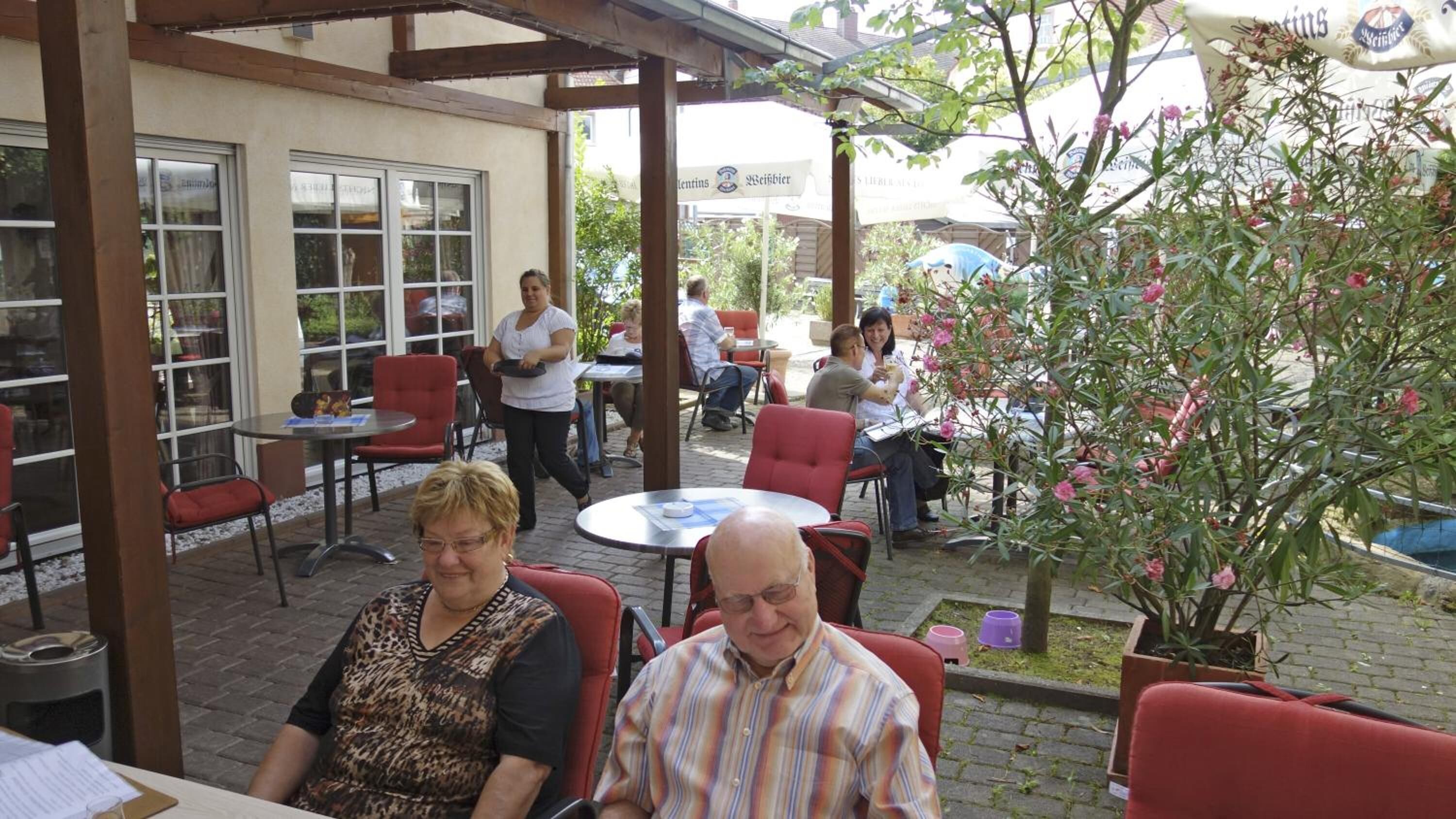 Un patio al aire libre con sillas y mesas rojas, una zona de estar cubierta y una planta en maceta con flores rosadas.