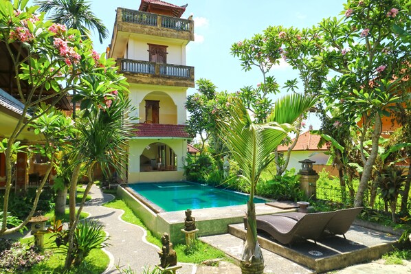 A pool area with a clear blue pool, surrounded by lush greenery and a multi-story building with balconies. - Ubud Kerta City Hotel (Ubud)