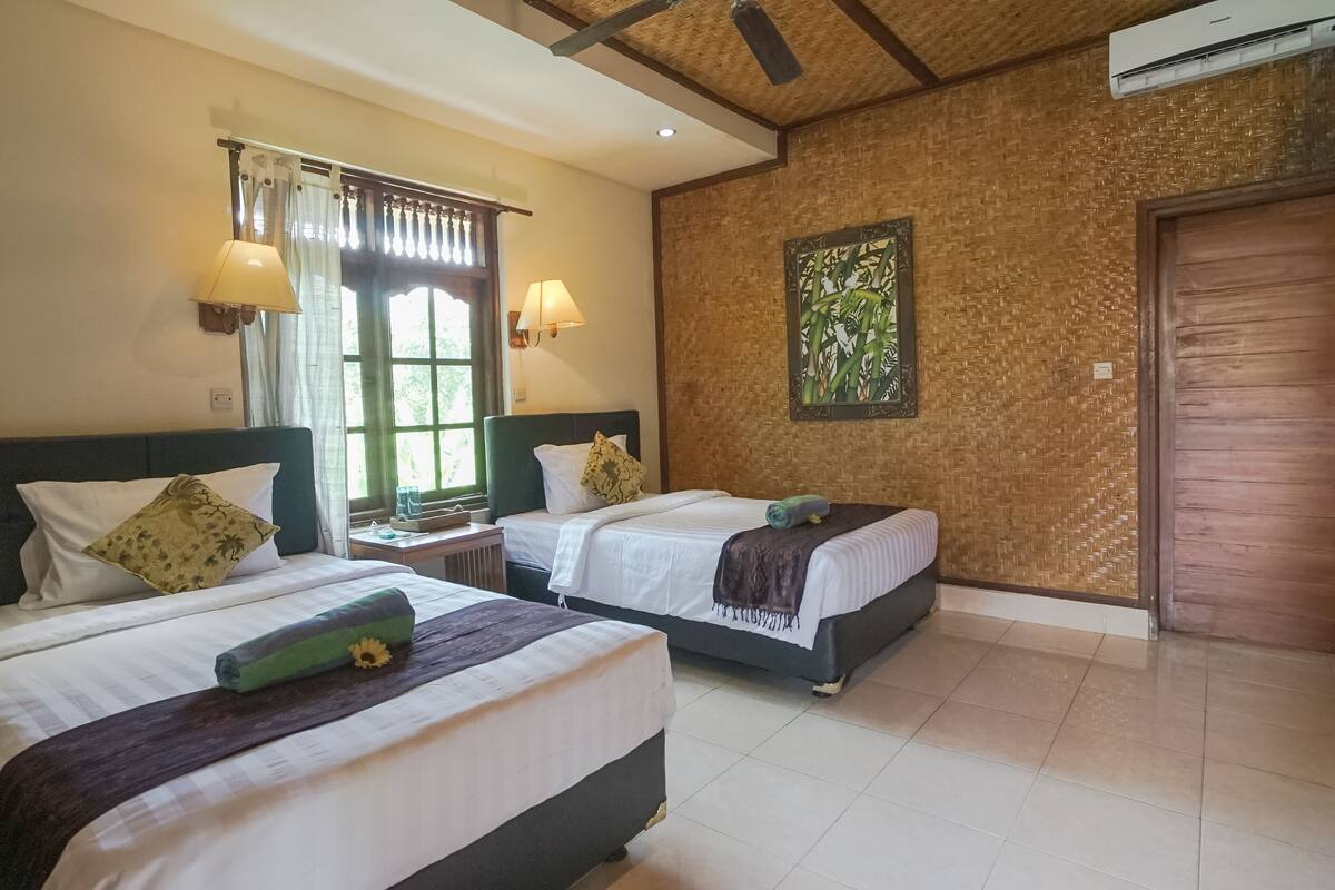 A hotel room with two beds, a window with curtains, a wall-mounted air conditioner, and a wooden door.