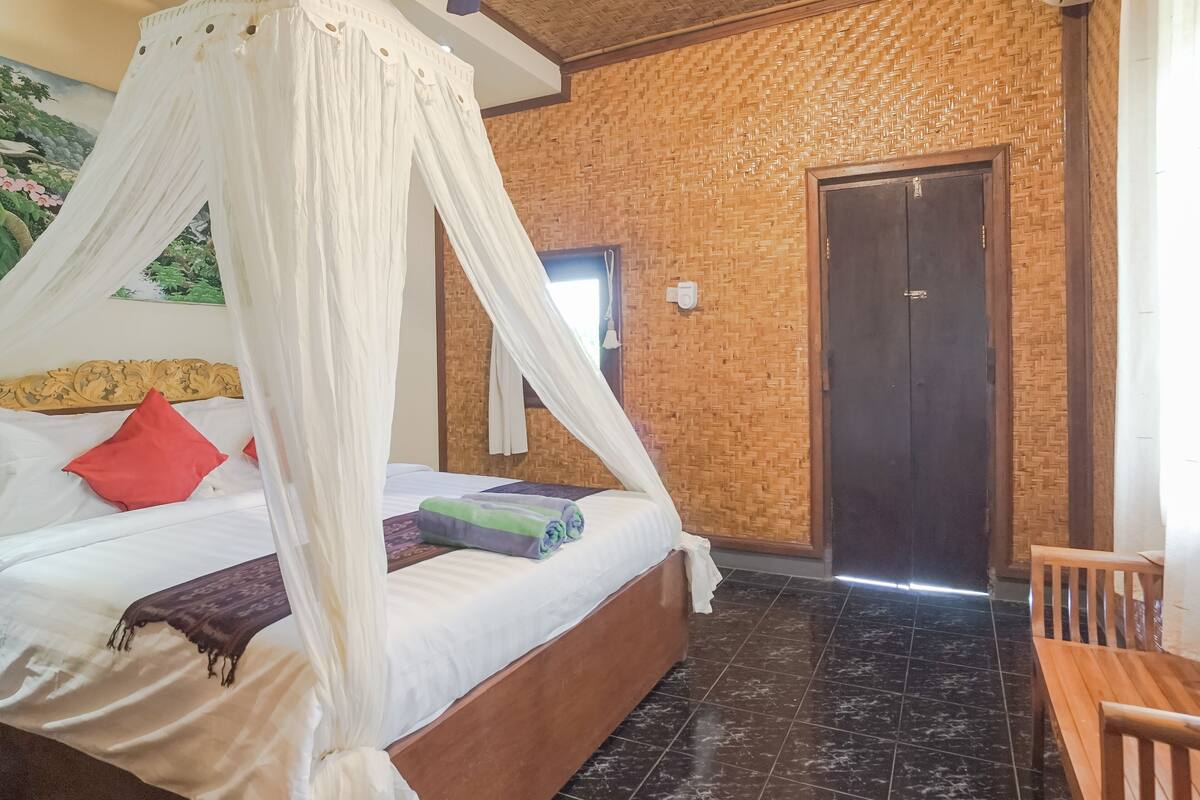 A room with a bed, a wooden bench, a door, and a wall with a woven pattern.