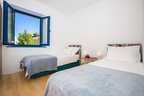 A bedroom with two beds, a window with blue frames, and a view of greenery outside.