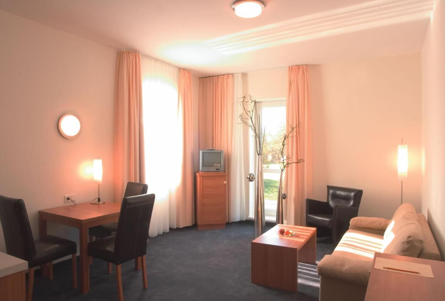 A hotel room with a dining table, two chairs, a sofa, a coffee table, a TV, and a vase with branches.