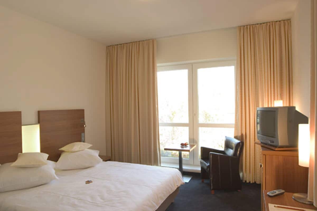 A hotel room with a bed, a TV on a wooden cabinet, a desk with a chair, and a window with curtains.