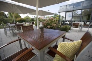Uma área de pátio ao ar livre com mesa e cadeiras de madeira, rodeada por guarda-sóis e plantas em vasos.