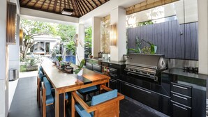A modern kitchen with a built-in grill, wooden dining table with blue chairs, and a view of the garden. - Sunset Paradise Villa Jimbaran 8008 (Kedonganan)