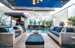 A modern outdoor lounge area with wicker furniture, a pool, and lush greenery. - Sunset Paradise Villa Temple Hill (Jimbaran)