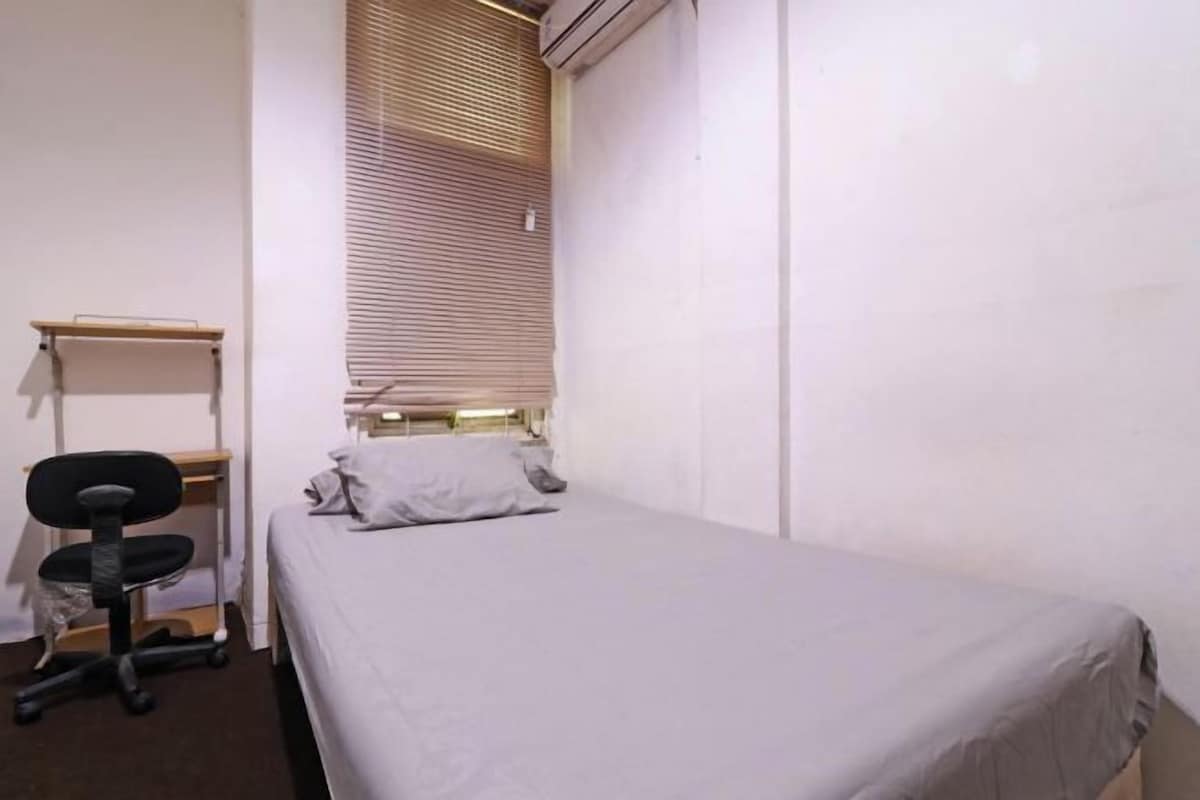 A small, single-bed room with a desk and chair, a window with blinds, and a wall-mounted shelf.