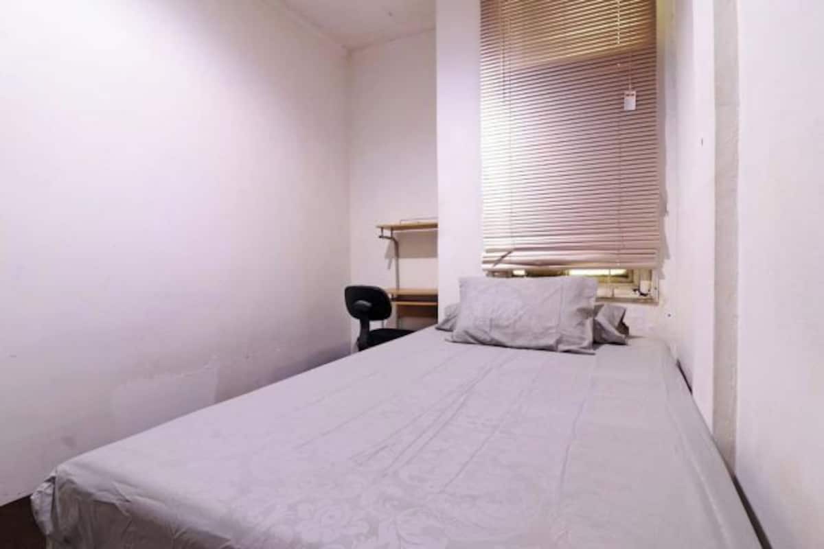 A small, single-bed room with a desk and chair, a window with blinds, and a white wall.