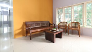 A room with a wooden bench, two wooden chairs, a small table with a plant, and a large window with a view of greenery. - Bedur Homestay Syariah (Bekasi)